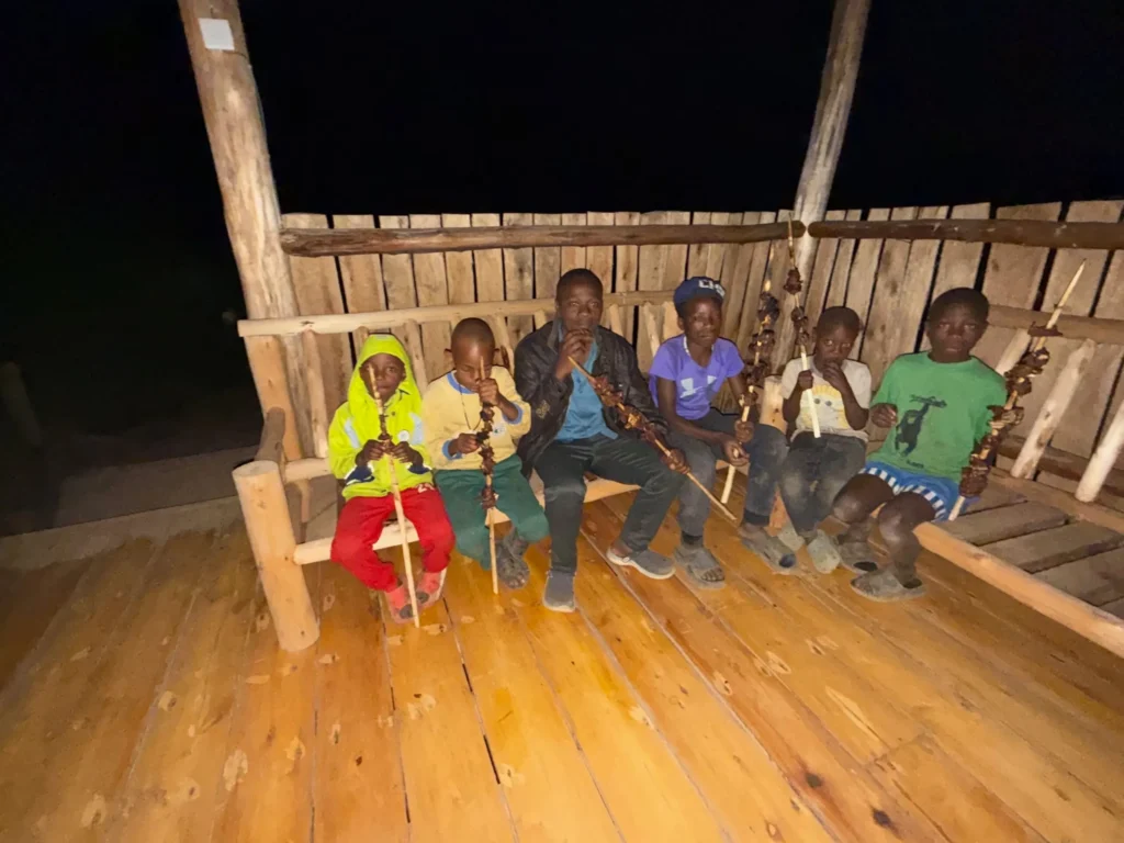 Six children enjoying barbecue skewers on wooden bench at night in rustic setting.