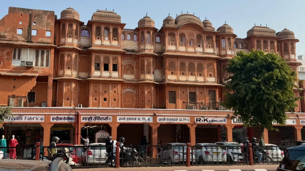 Historic Jaipur building with red sandstone facade, domes, shops, motorbikes, sunny day.