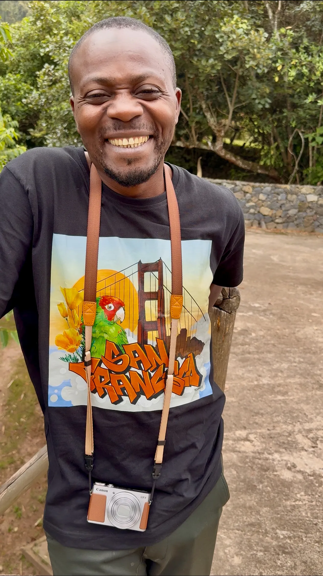 Smiling man wearing black graphic t-shirt with parrot artwork, leaning on railing, camera around neck.