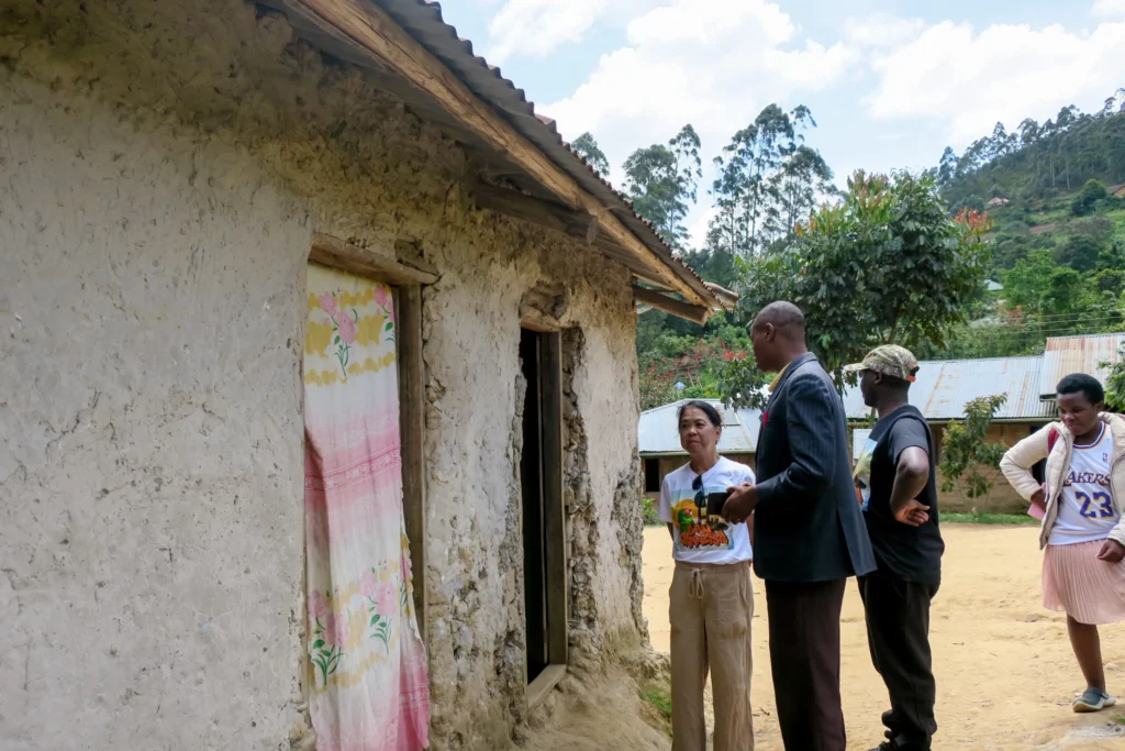 Group of people outside rustic mud house with curtain, hills in background.