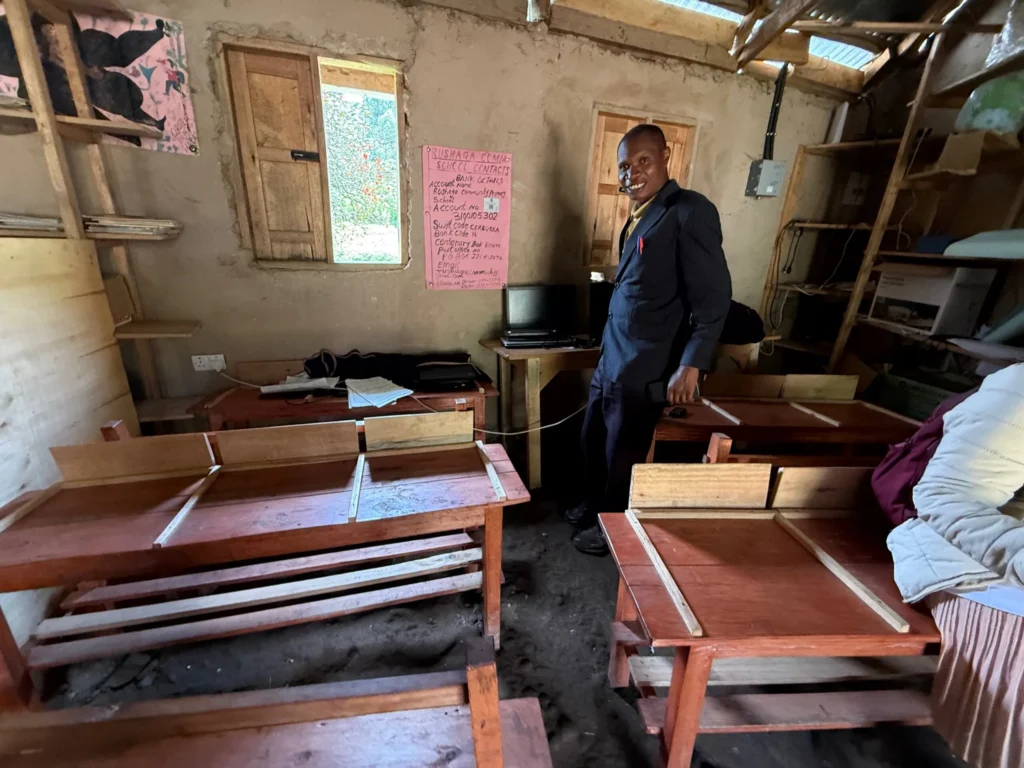 Smiling man in warm classroom with wooden desks, pink poster, small window.