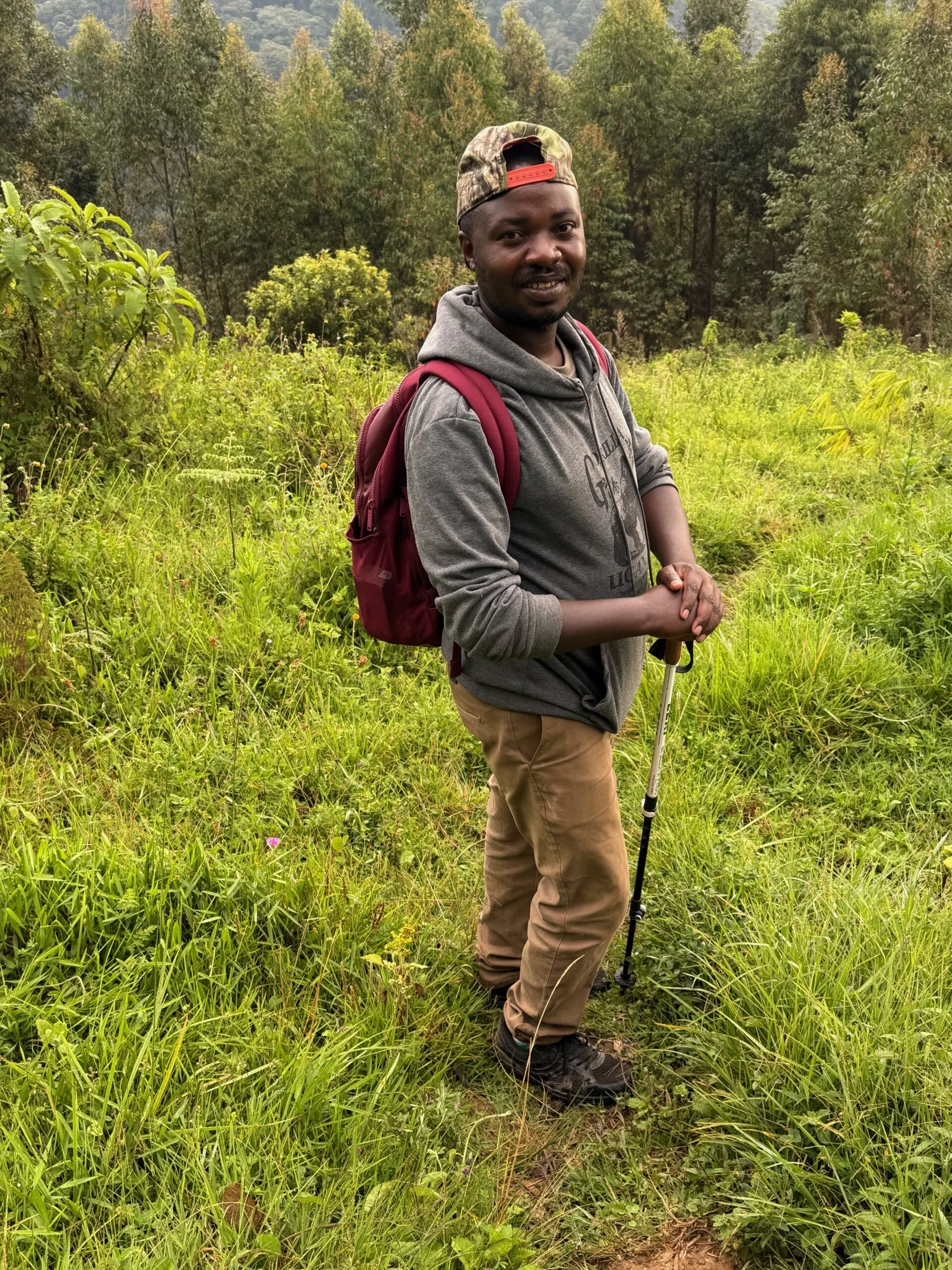 Smiling hiker in gray hoodie and cap on green forested hillside.