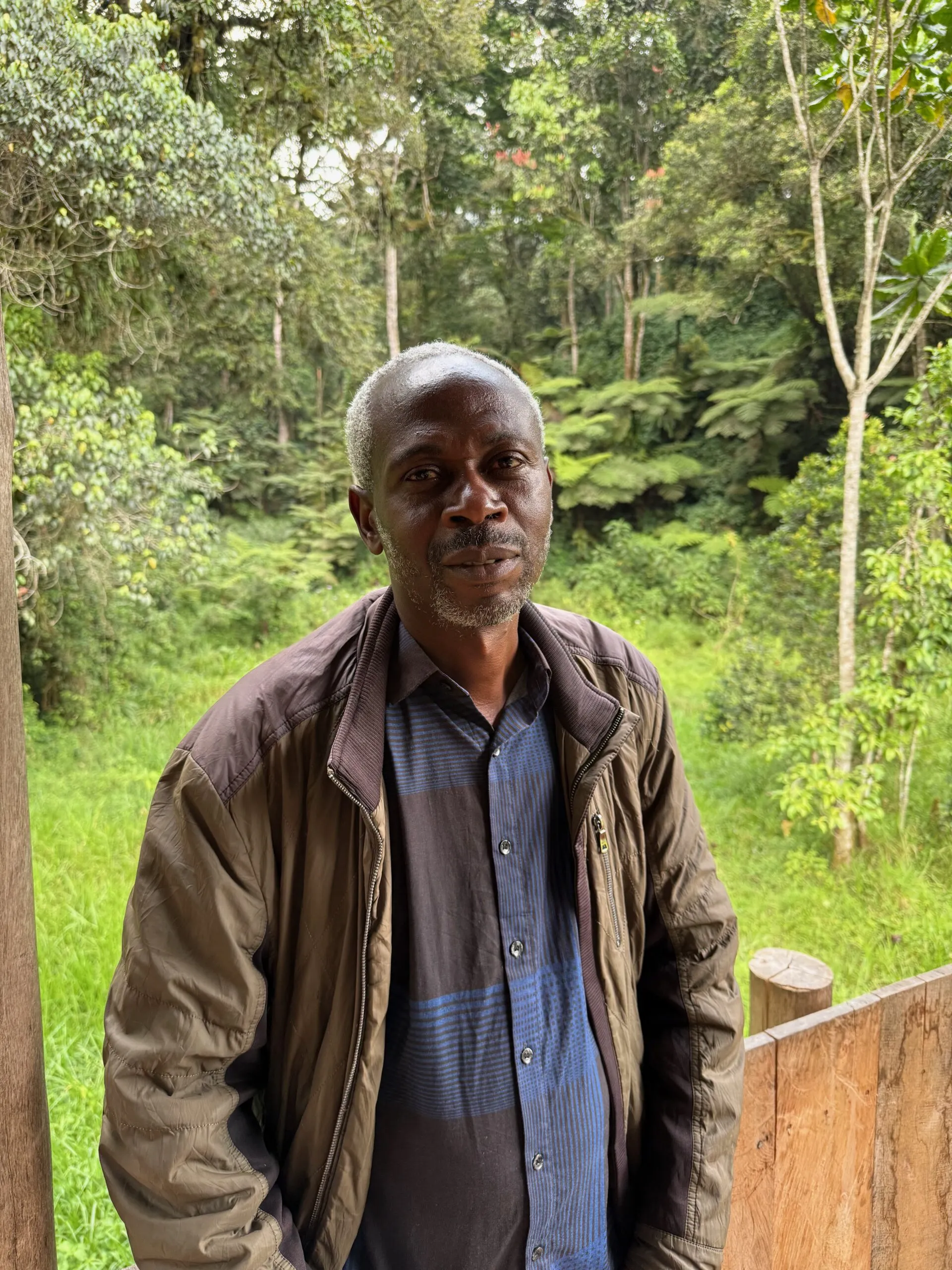 Man in brown jacket standing peacefully in dense green forest with trees.
