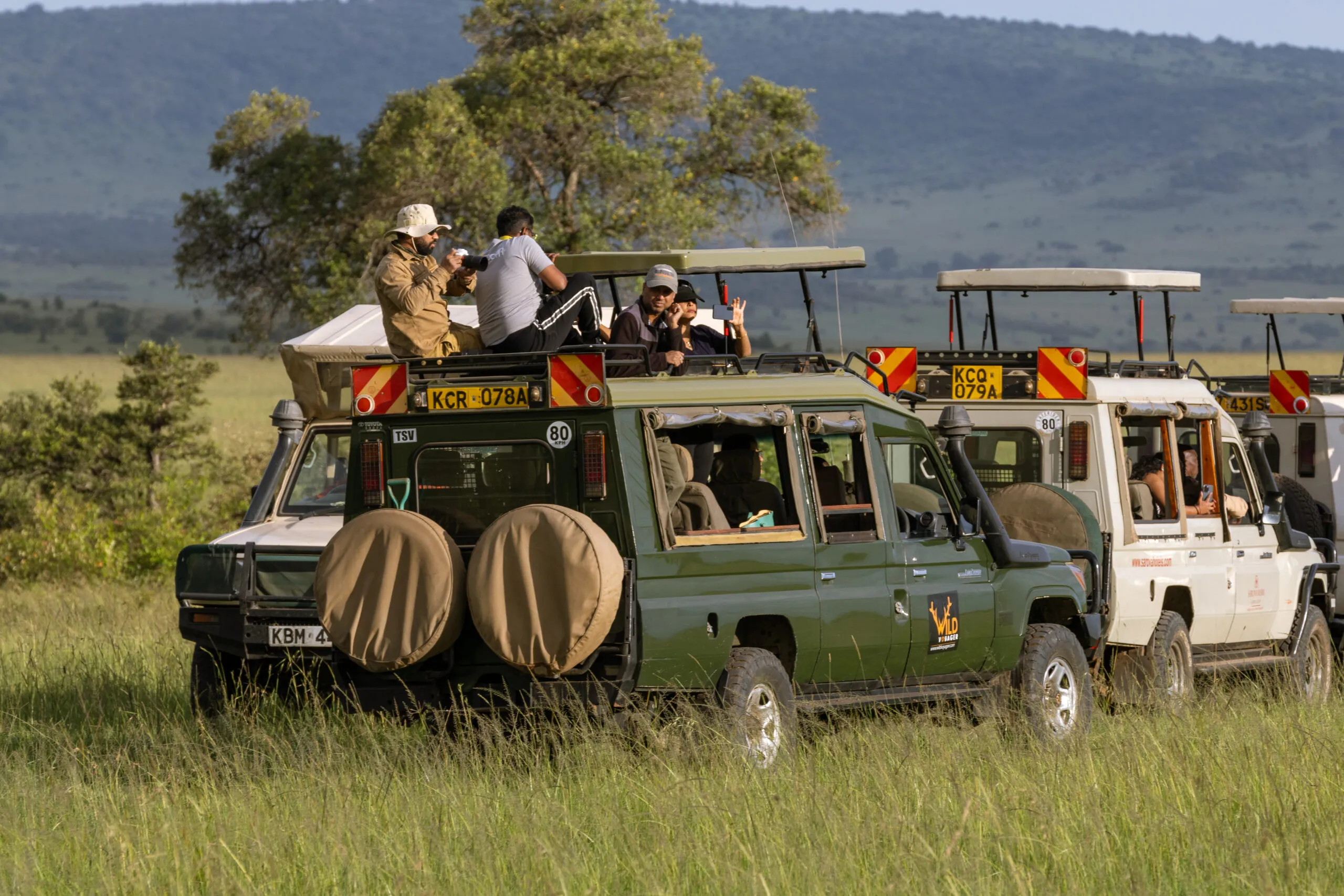 Tourists in safari vehicles photographing vibrant green savanna with distant hills adventure.