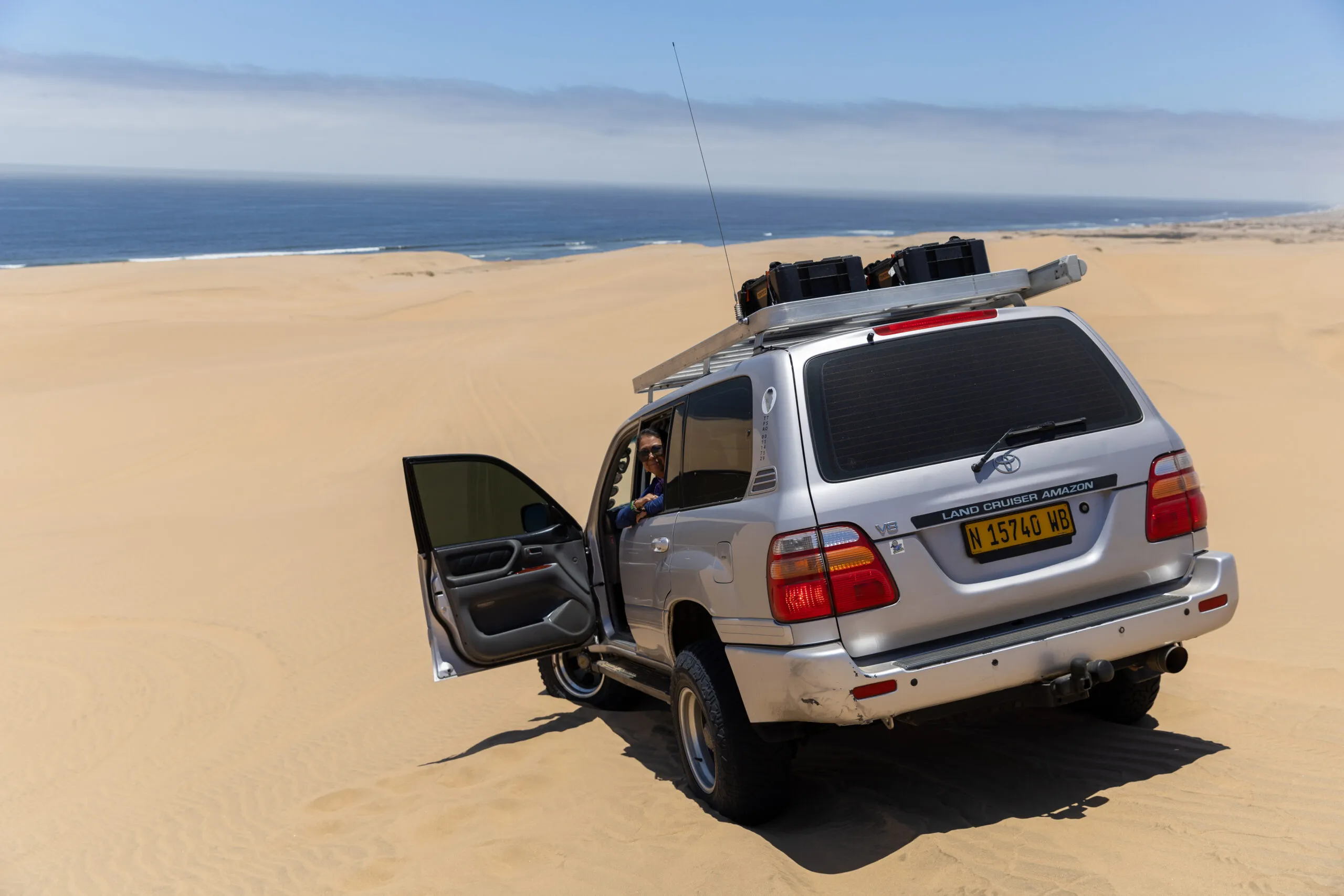 Silver SUV on desert dune with open door, ocean in background adventure.