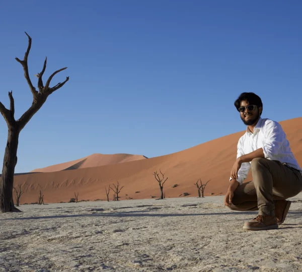 Man kneeling on cracked desert ground near leafless tree and dunes.